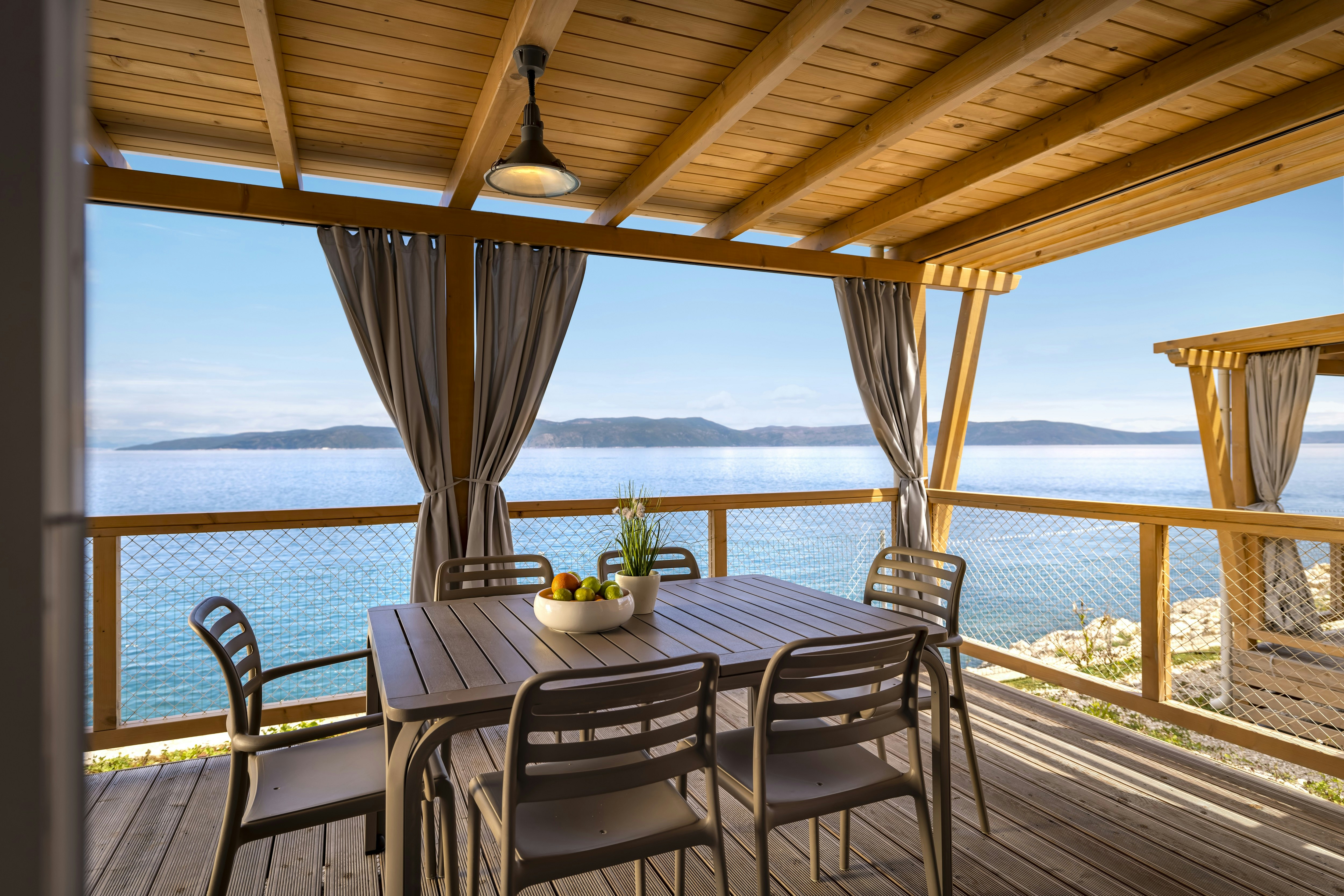 Holzterrasse mit Tisch und Stühlen, Vorhänge am Pavillon und Seeblick durch das Geländer.