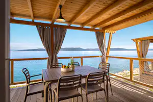 Holzterrasse mit Tisch und Stühlen, Vorhänge am Pavillon und Seeblick durch das Geländer.