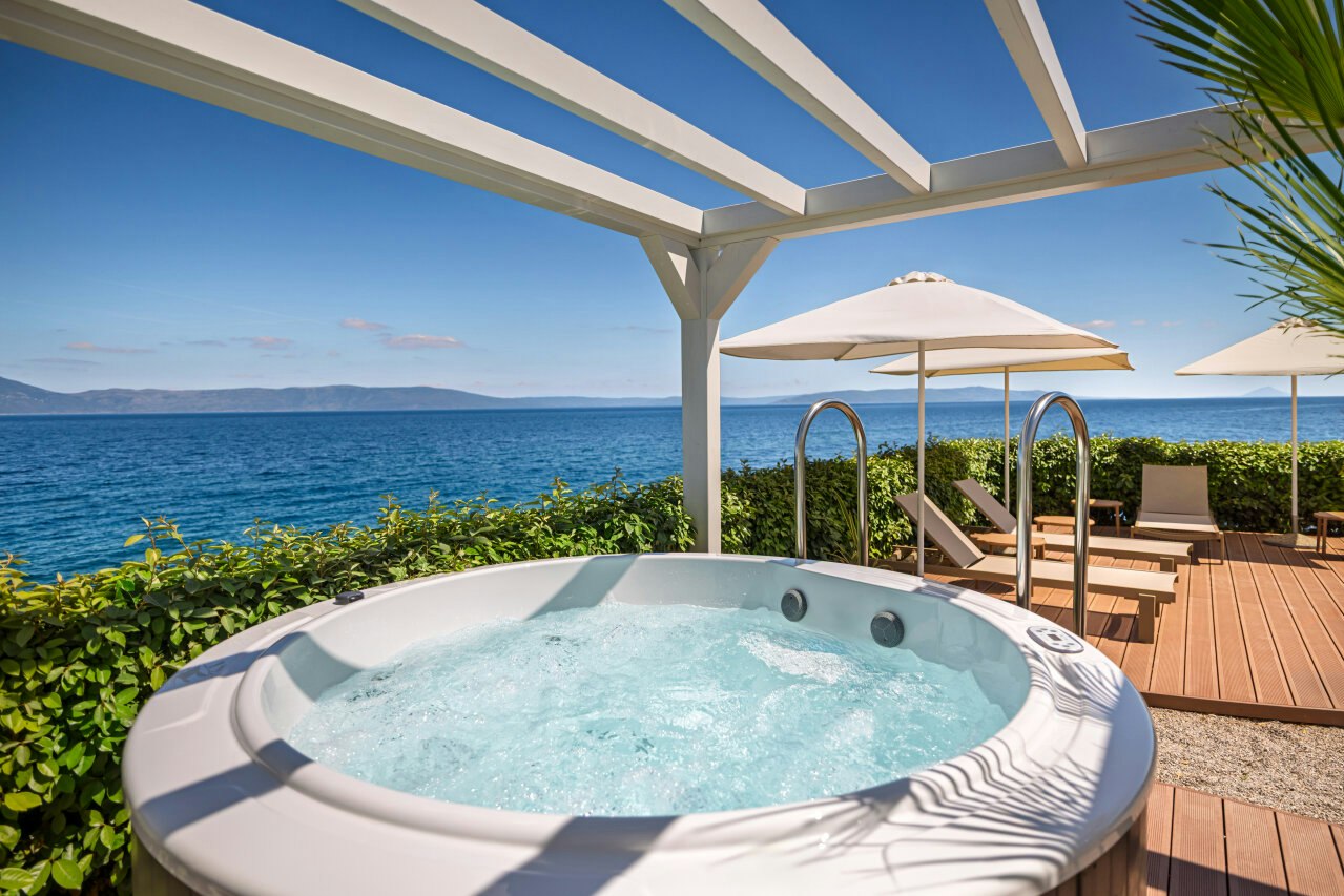Whirlpool auf einer Holzterrasse mit Sonnenschirmen, Blick auf das Meer und die Küste, im Hintergrund Berge.
