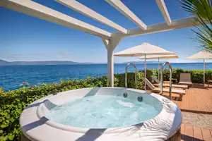 Whirlpool auf einer Holzterrasse mit Sonnenschirmen, Blick auf das Meer und die Küste, im Hintergrund Berge.