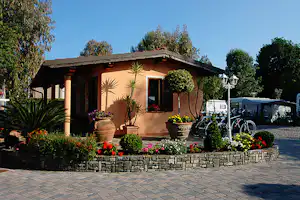 Bungalow mit Veranda, Blumenbeeten und Pflasterweg, im Hintergrund Bäume und Fahrräder am Stellplatz.