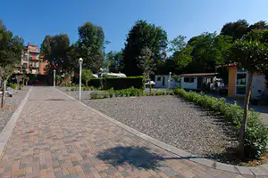 Gepflasterter Weg auf dem Campingplatz, daneben Kiesfläche und mehrere Wohnwagen, Hecken und Bäume am Rand.