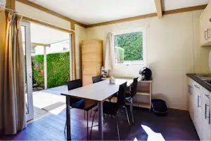 Wohnbereich eines Mobilheims mit Esstisch, Stühlen und Küchenzeile, große Fenster zur Terrasse und Blick in den Garten.