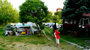 Wohnwagen und Campingbusse auf einem Rasenstellplatz, dazwischen Bäume und ein Weg zum Gebäude.