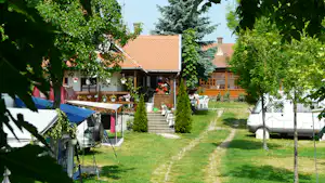 Wohnwagen und Zelte auf einem Campingplatz, dazwischen ein Kiesweg und Rasen, umgeben von Bäumen und Gebäuden.