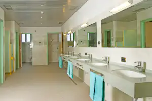 Gemeinschaftsbad mit langen Waschbecken, Wasserhähnen und grünen Türen, Handtüchern am Spülbecken und Fliesenboden.