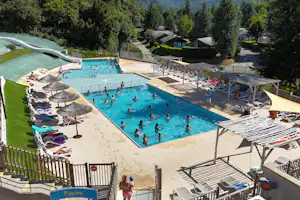 Camping-Pool mit vielen Liegen, Sonnenschirmen und Menschen im Wasser, umgeben von Hecken und Bungalows im Hintergrund.
