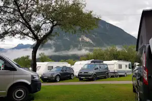 Stellplätze auf einem Campingplatz mit Wohnmobilen und Autos, Rasenfläche, Baum und Berge im Hintergrund.