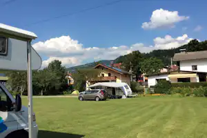 Wohnwagen auf einem Stellplatz mit grünem Rasen, daneben ein Auto und ein weiteres Wohnmobil vor Häusern und Bäumen.