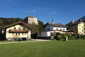 Campingplatz mit Rasenfläche, Wohngebäuden und Terrasse, im Hintergrund ein steinernes Schloss und Bäume unter blauem Himmel.