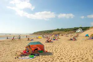 Camping am Strand: Zelt, Picknickdecke und Sonnenliegen auf dem Sand, daneben weitere Strandgäste und ein Küstenabschnitt.