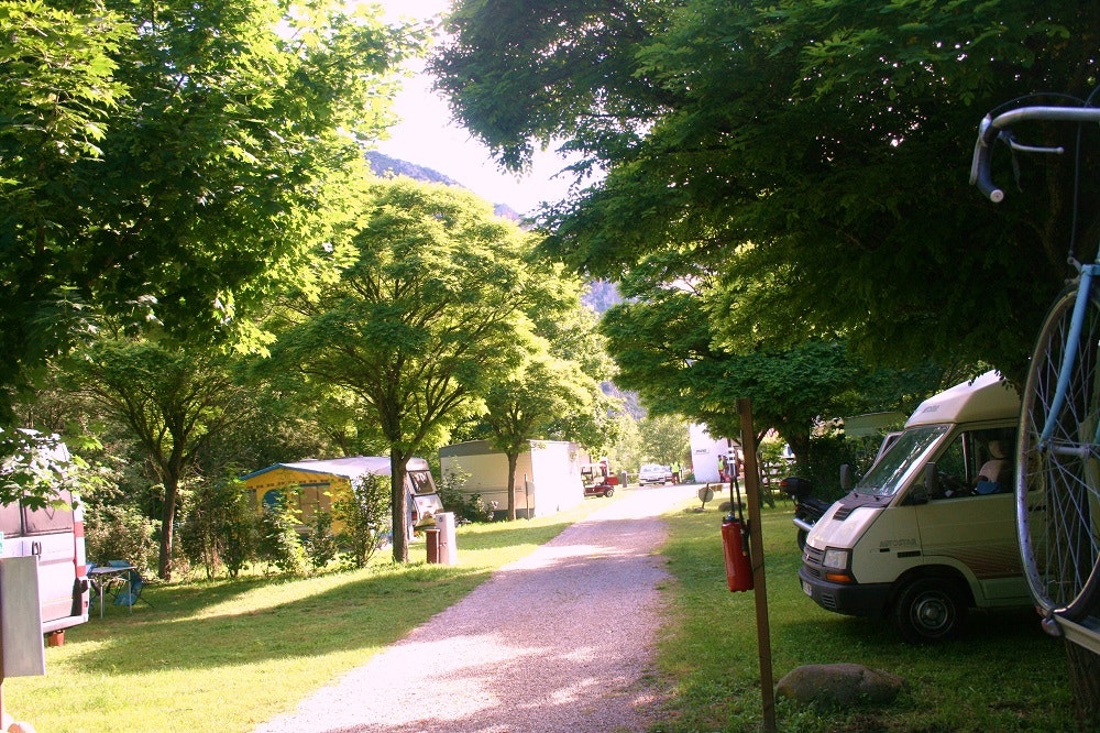 Campingweg zwischen Bäumen, links Wohnwagen und rechts ein geparktes Fahrzeug am Stellplatz.