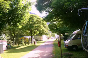 Campingweg zwischen Bäumen, links Wohnwagen und rechts ein geparktes Fahrzeug am Stellplatz.