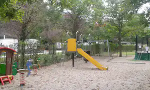 Spielplatz am Campingplatz mit gelber Rutsche, Klettergerüst und Sandboden, umgeben von Bäumen und Zäunen.