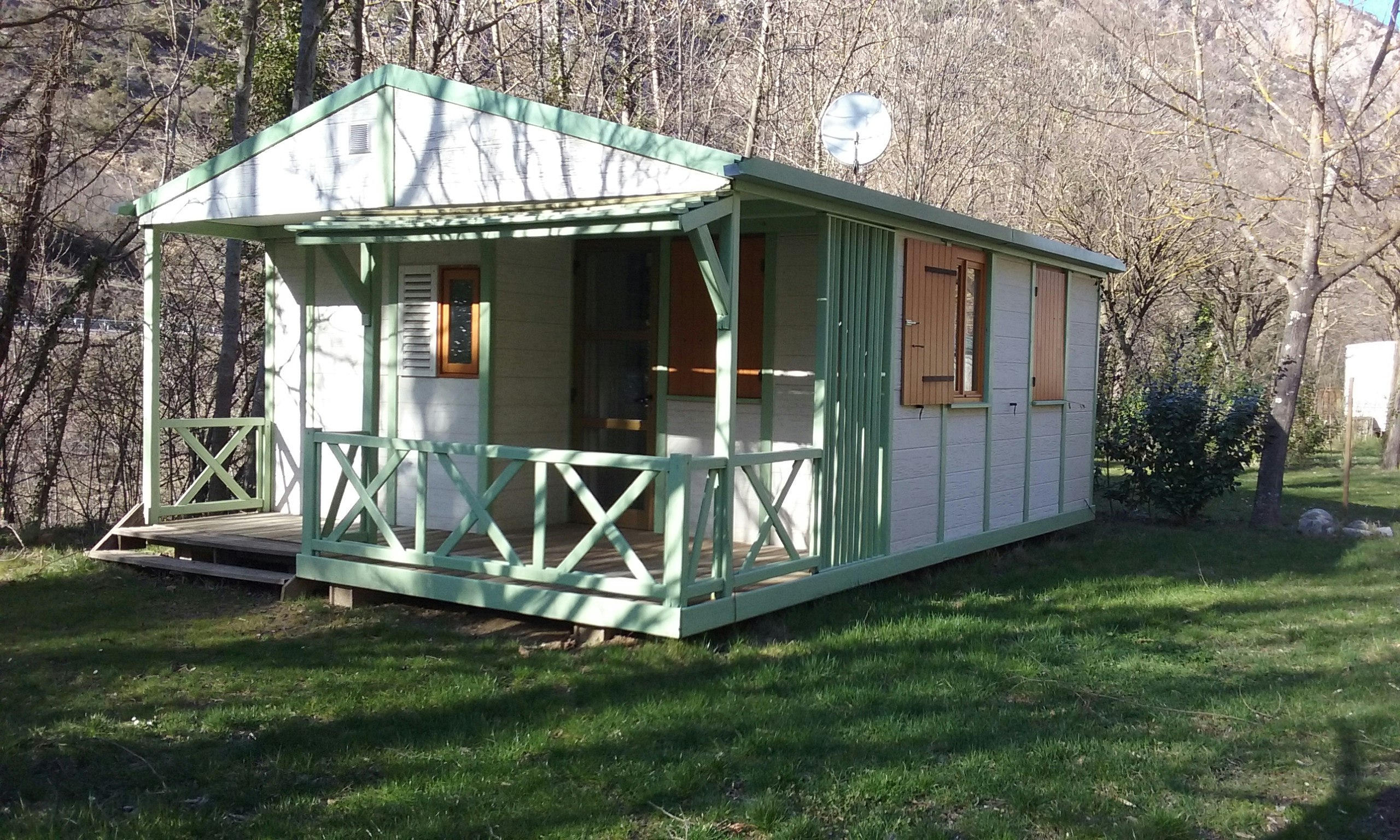 Grünes Mobilheim mit Veranda auf Holzpodest, umgeben von Bäumen und Rasenfläche am Campingplatz.