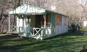 Grünes Mobilheim mit Veranda auf Holzpodest, umgeben von Bäumen und Rasenfläche am Campingplatz.