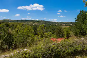 Weite Aussicht über grüne Hügel und Wälder, im Vordergrund Büsche und Steine, im Hintergrund Häuser mit roten Dächern.