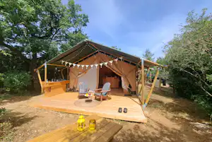 Zeltlodge mit Terrasse, zwei Stühlen und Holztisch, Lichterkette am Dach, umgeben von Bäumen im Campingbereich.