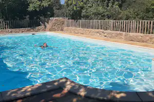 Schwimmbecken mit klarem Wasser, Person im Pool, Steinmauer und Holzsichtschutz im Campinggarten.