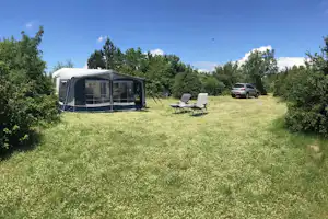 Wohnwagen mit Vorzelt auf einer grünen Wiese, daneben zwei Campingstühle und ein Auto am Stellplatz.