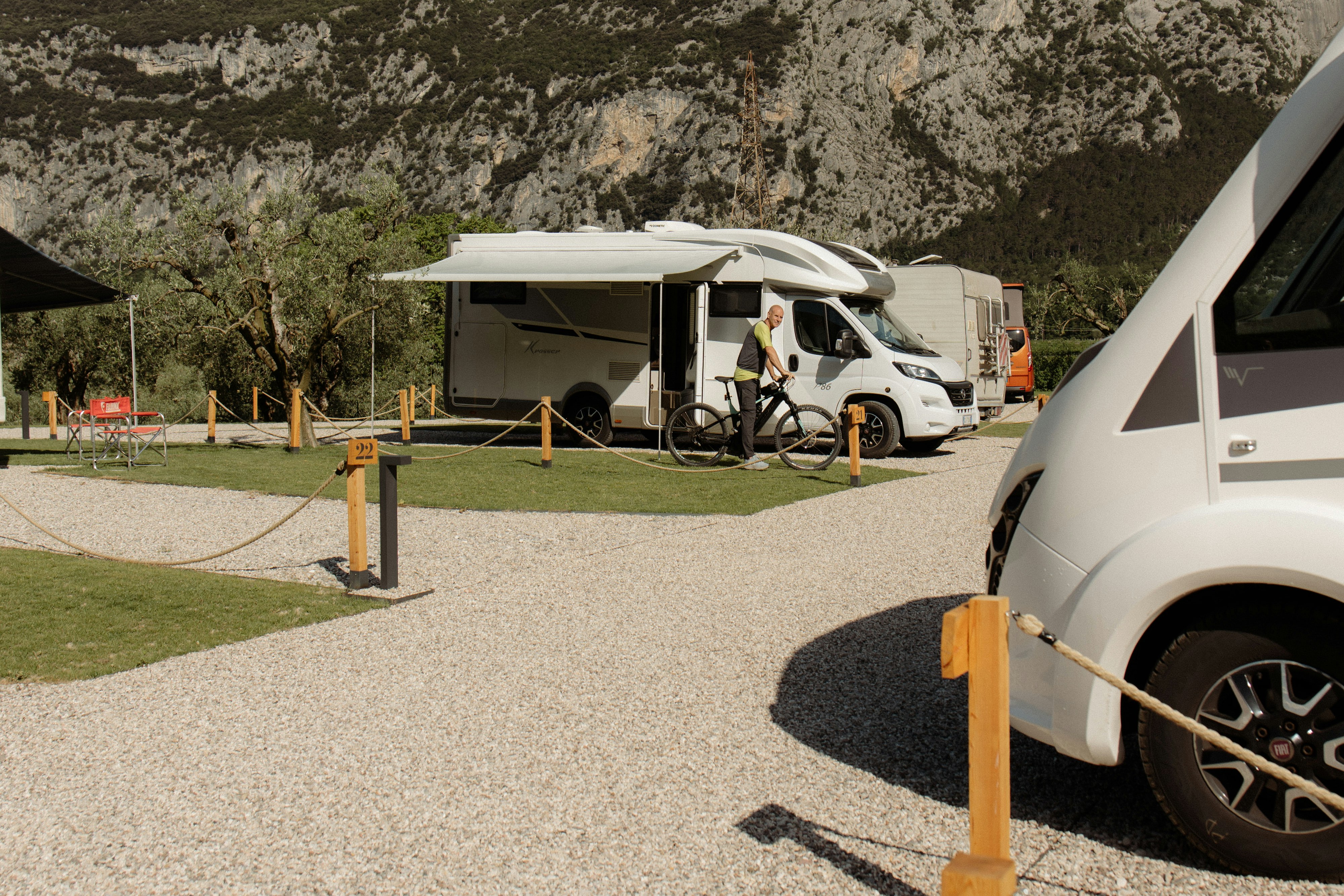 Wohnmobil auf einem Stellplatz mit Kiesfläche, Holzpfosten und Blick auf einen felsigen Berg im Hintergrund.