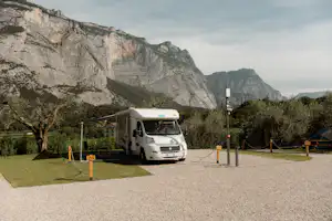 Wohnmobil auf einem Kiesstellplatz, im Hintergrund Berge und Felsen, daneben Bäume und ein Stromanschluss.