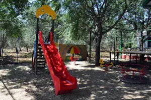 Rote Rutsche auf einem Spielplatz mit Sitzbank und Tisch, umgeben von Bäumen und Schatten im Campingbereich.