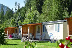 Wohnwagen mit Holzveranda und Sitzgruppe auf dem Campingplatz, umgeben von hohen Bäumen und grüner Wiese.