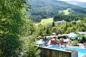 Camping-Pool mit Liegen und bunten Zelten, umgeben von grünen Bäumen und Bergen im Hintergrund.