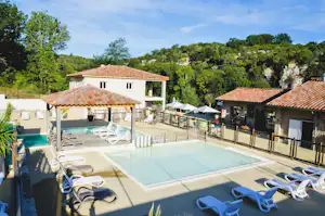 Großer Camping-Pool mit Liegen und Sonnenschirmen, dahinter weiße Gebäude und Bäume im grünen Hintergrund.