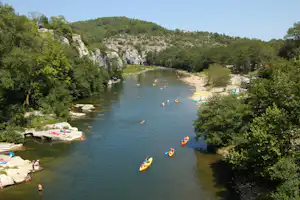 Fluss mit mehreren Kajaks, Ufer mit Booten und Steinen, umgeben von Bäumen und bewaldeten Hügeln.