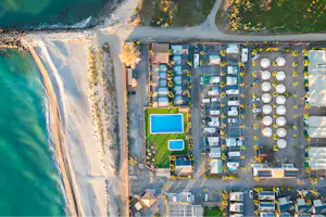 Luftaufnahme eines Campingplatzes mit Stellplätzen, Wohnmobilen und Zelten am Strand, umgeben von Wegen und Bäumen.