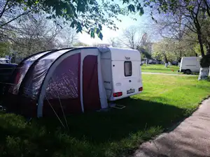 Zelt auf einem Campingplatz-Stellplatz neben einem Wohnwagen, Rasenfläche und Weg, Bäume im Hintergrund.