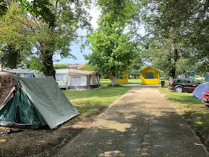 Campingzelt und Wohnwagen auf einer Wiese, gepflasterter Weg, gelbe Hütte und viele Bäume im Hintergrund.