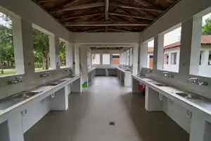 Gemeinschaftsküche mit langen Spülbecken, Wasserhähnen und Fliesenboden unter Holzdecke, im Hintergrund weitere Waschbereiche.