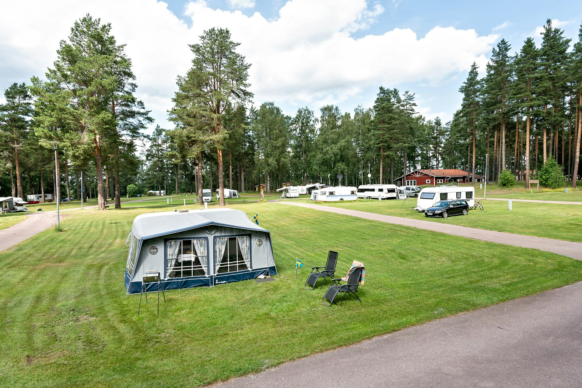 Campingplatz mit Wohnwagen und Vorzelt auf dem Rasen, daneben Campingstühle und ein Weg zwischen den Stellplätzen.