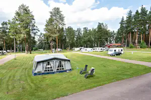 Campingplatz mit Wohnwagen und Vorzelt auf dem Rasen, daneben Campingstühle und ein Weg zwischen den Stellplätzen.