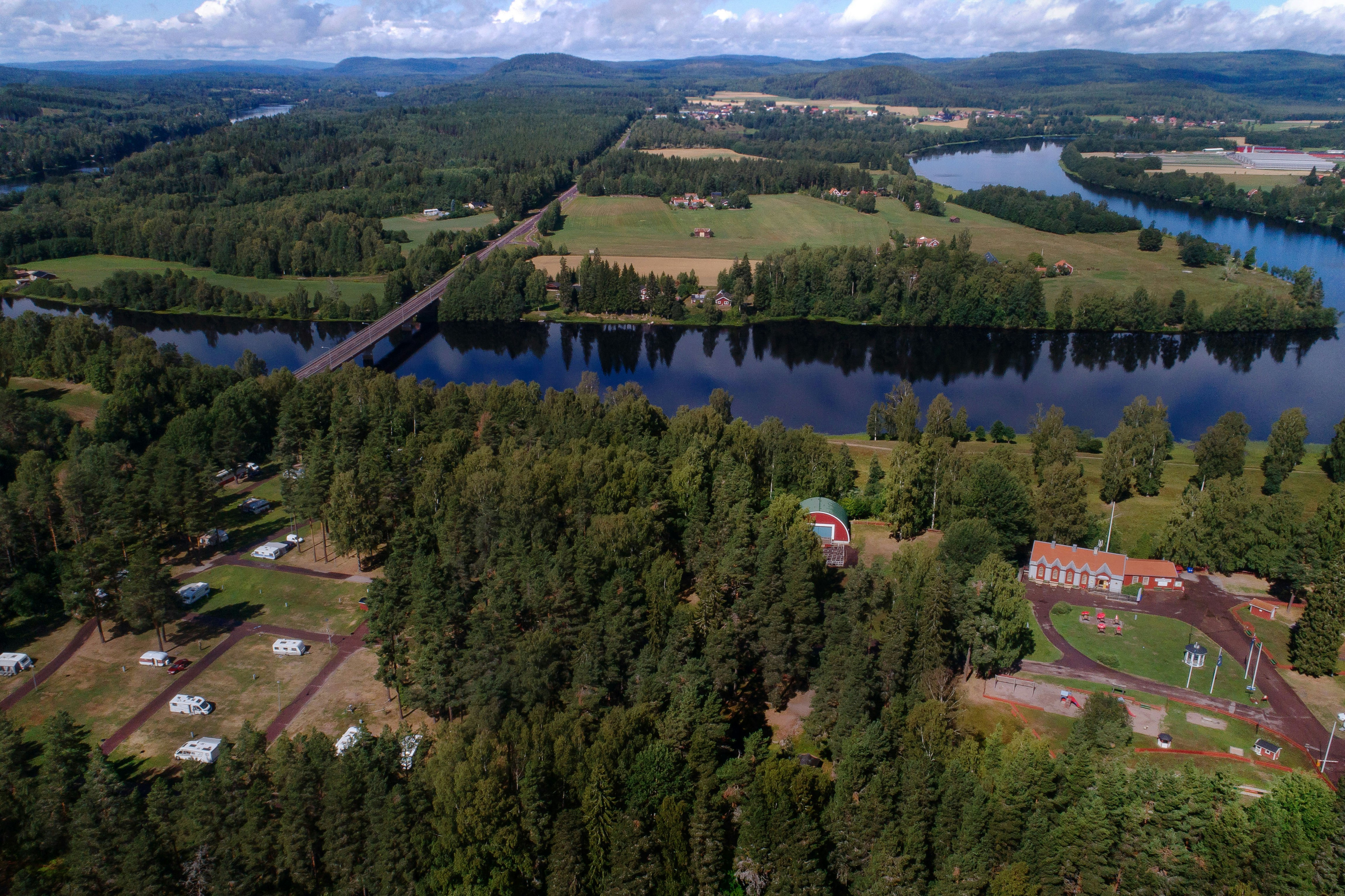 Luftaufnahme eines Campingplatzes am See mit Stellplätzen, Wegen, Bäumen und einem kleinen Gebäude am Ufer.