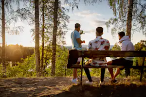 Familie am Camping-Tisch im Wald, Erwachsene und Kinder sitzen und schauen in die Ferne bei goldenem Sonnenlicht.