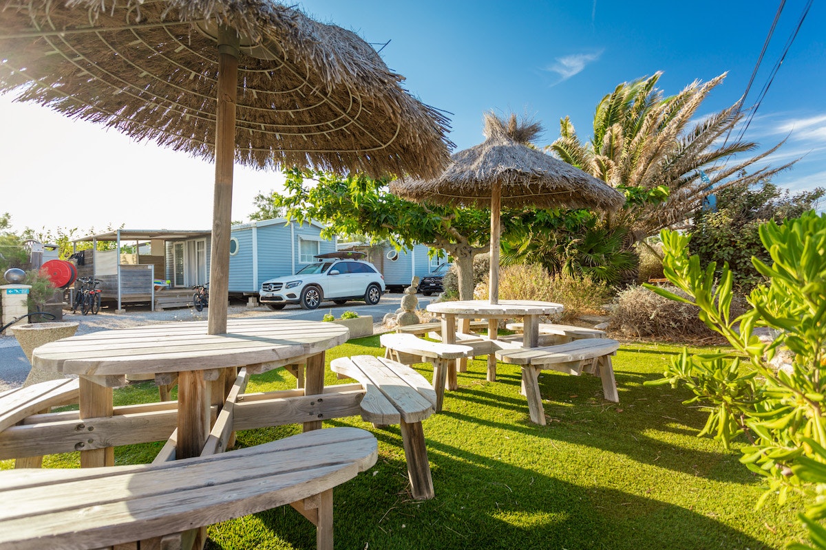 Holztische und Bänke unter Strohdach auf einer Camping-Terrasse, daneben Rasen und Palmen im Garten.