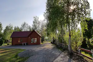 Holzhaus am Campingplatz mit Eingangstür, Bank und Weg, umgeben von Birken und Rasen im grünen Gelände.