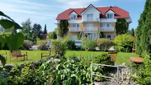 Campinghaus mit roten Ziegeldach, Balkonen und Fenstern, umgeben von Gartenbeeten, Rasen und mehreren geparkten Autos.