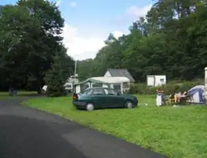 Grüner Campingplatz mit geparktem Auto auf dem Stellplatz, Zelt im Hintergrund und Bäume entlang des Weges.