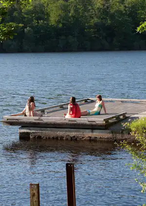 Campingsteg am See mit drei Personen auf einer Holzplattform, umgeben von Bäumen und ruhigem Wasser.