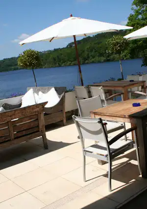 Terrasse am See mit Holztisch, Stühlen und Sonnenschirmen, im Hintergrund grüner Wald und ruhiges Wasser.