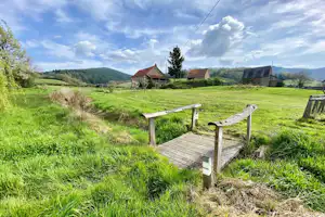 Holzsteg und Bank auf einer grünen Campingwiese, im Hintergrund Bauernhäuser, Weiden und Hügel im Tal.