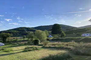 Campingwiese mit Wohnwagen auf dem Stellplatz, umgeben von Bäumen und Hügeln unter blauem Himmel.