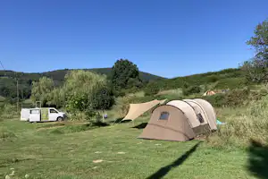 Campingzelt auf einer Wiese, daneben ein weißer Camper-Van und weitere Zelte vor grünen Hügeln und Bäumen.