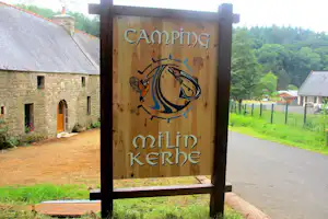 Holzschild mit Aufschrift „Camping Amilin Kerhe“ vor einem Campingplatz mit Steingebäuden und Wiese.