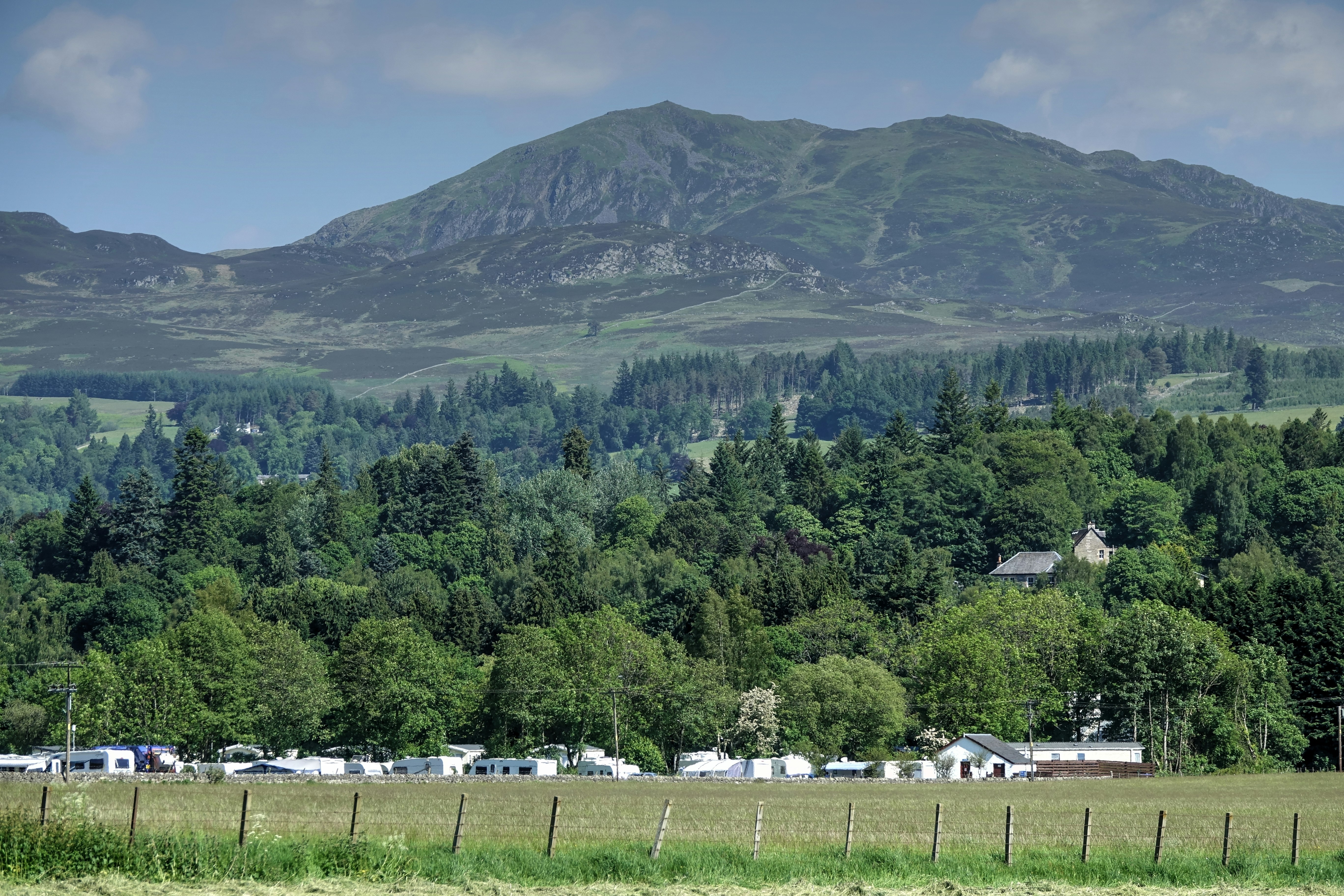 Campingplatz mit Wohnwagen und Zelten am Rand einer Wiese, Blick ins Tal mit Wald und hohen Bergen.